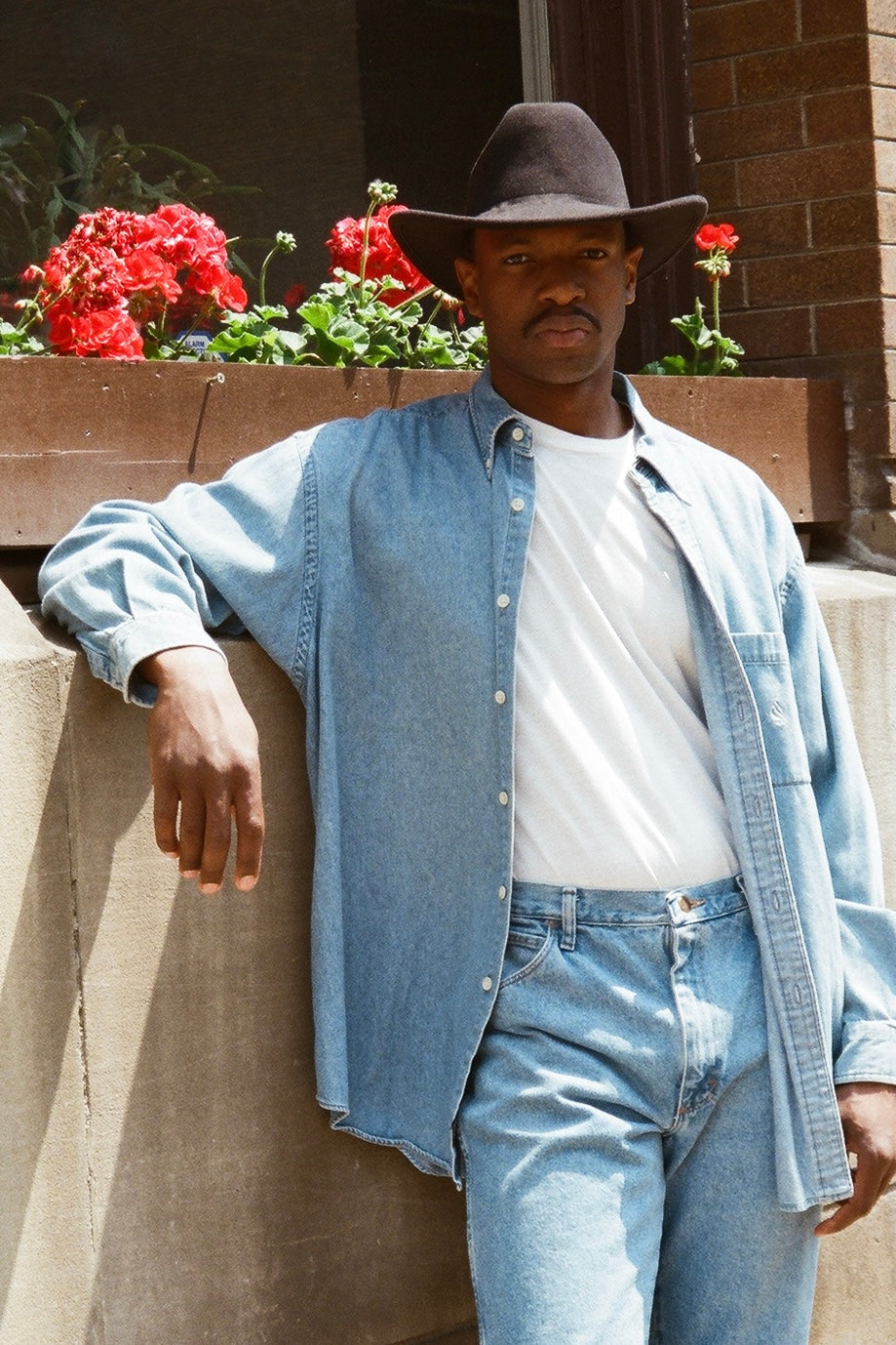 Man wearing a denim shirt, white t-shirt, and jeans standing outdoors with flowers in the background.